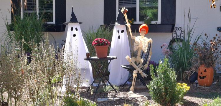 skeleton and ghost sitting at a cafe table with coffee, standing ghost next to them in a front yard
